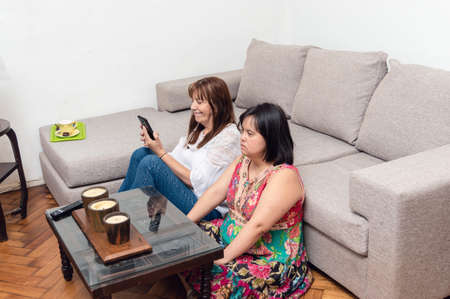 overhead view caucasian latin mom and daughter sitting on living room floor using phone and watching...