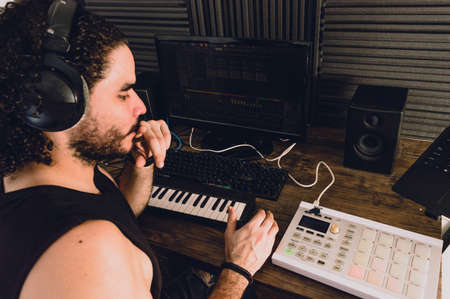 young latin caucasian man in his home studio, with keyboards, computer and midi controller, thinking and producing music, music industry concept.の写真素材