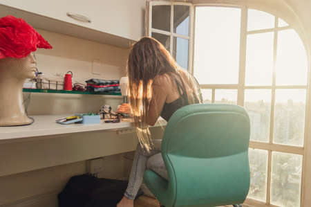 young latin caucasian woman by her window using her sewing machine in her sewing studio, sewing...