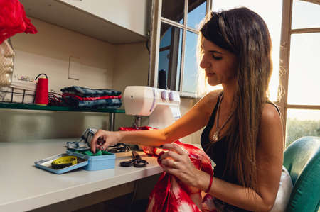 Side view of pretty Argentinian Caucasian young woman smiling and working sitting in her sewing workshop, with sewing tools and sewing machine on the table.の写真素材