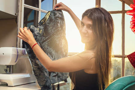 latin caucasian young woman sitting smiling and looking at the camera holding a black fabric in her sewing workshop, with a sewing machine on the table, manufacturing and designing fashion clothes.の写真素材