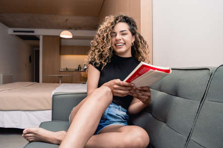 young caucasian girl sitting on the sofa in her apartment with the kitchen and her bed behind her, holding a book happily smiling looking at the camera.の写真素材