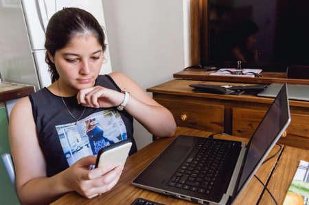 Caucasian young woman in her home office checking messages on the phone while working on the...