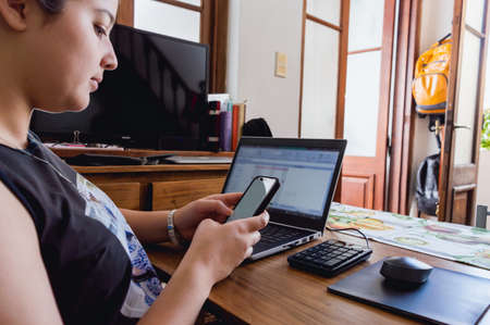 back view of enterprising young caucasian girl in home office checking her phone while working with computer on table and front door in backgroundの写真素材