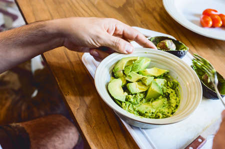 seated caucasian man holding with his hand on the table a white porcelain bowl with cut and crushed avocado.の写真素材