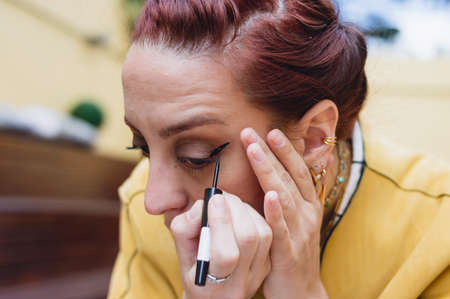 close-up of caucasian red hair young woman touching black eyeliner on her face, staring at mirror, applying makeup at home.の写真素材