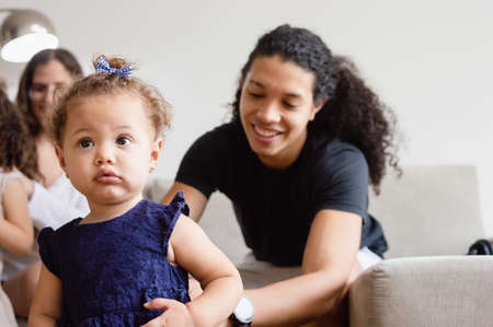 Caucasian baby in blue dress standing in the living room at home looking to the side with her blurred father sitting on the sofa smiling behind her with his wife.の写真素材