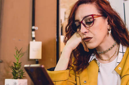 young redhead caucasian woman inside a restaurant is serious and concentrated reading bad news on her phoneの写真素材