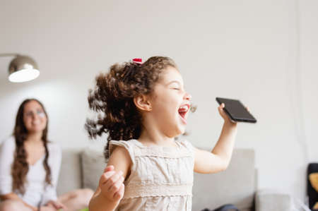 little caucasian girl with curlers jumping, with closed eyes, playing and laughing in the living room at home with a phone in her handの写真素材