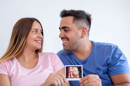 portrait of happy pregnant heterosexual caucasian young couple laughing inside bedroom looking at each other and showing ultrasound printout of their babyの写真素材