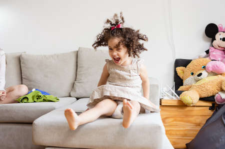 little caucasian girl with curlers in diapers happy screaming jumping on the living room sofaの写真素材