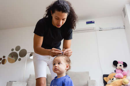 young latin caucasian father in the living room at home combing his little daughter with curls, he puts a blue clip on her hair, father's day and lifestyle concept.の写真素材