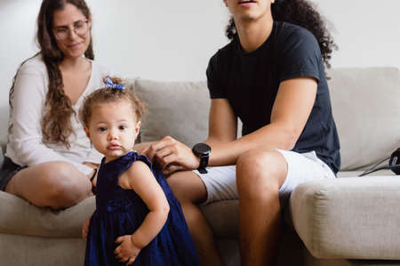 little caucasian baby standing in the living room looking at the camera while her dad finishes dressing her in a blue dress to go out, family and father's day concept, copy space.の写真素材