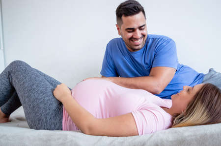 couple of caucasian hispanic latin venezuelan pregnant woman and man are smiling lying on the bed seeing each other in the bedroom, lifestyle and obstetrics concept.の写真素材
