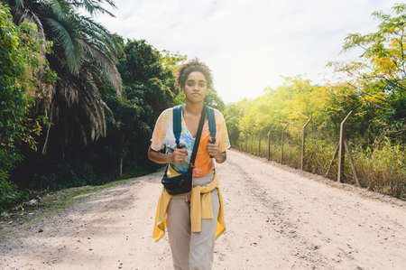 young brunette afro adventurous tourist girl walking in the ecological reserve park with a backpack, with the sky and trees behind her, looking at the cameraの写真素材