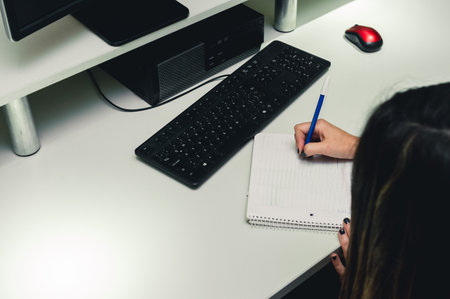 unrecognizable young woman sitting in her office writing in pad, with keyboard and computer cassette...
