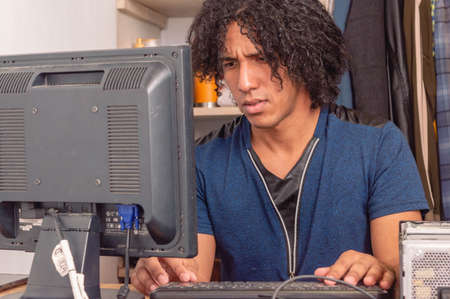young brunette man, with curlers, sitting indoors in the room, working on the computer.