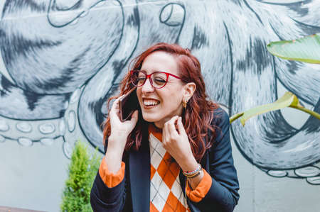 pretty caucasian redhead woman, wearing casual clothes and glasses, outdoors smiling and talking on the phone.の写真素材