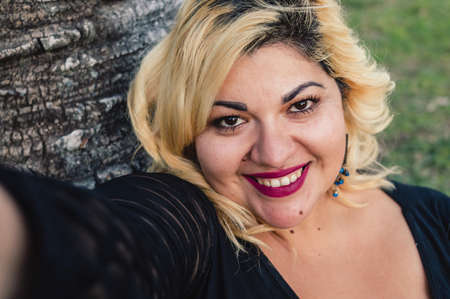 close up portrait beautiful blonde plus size caucasian young woman smiling taking a selfie outdoors sitting on grass, looking at camera, lifestyle concept.の写真素材