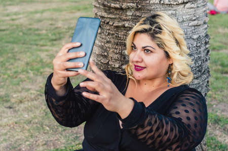 pretty plus size caucasian woman with blonde hair sitting outdoors in the park, leaning against a palm tree, taking a selfie with her phoneの写真素材