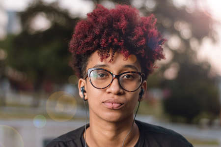 close up portrait outdoors beautiful young latin brunette woman with dyed red afro, glasses and headphones, serious looking at the cameraの写真素材