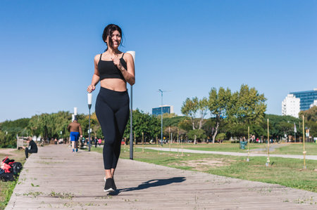front view latin caucasian woman wearing black sportswear, training, jogging and smiling outdoors in the morning, on a wooden walkway in the park, blue sky behind her with copy space.の写真素材