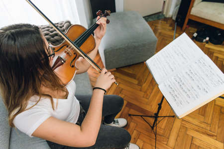 top view young caucasian woman sitting on sofa in living room at home practicing violin reading sheet music.の写真素材