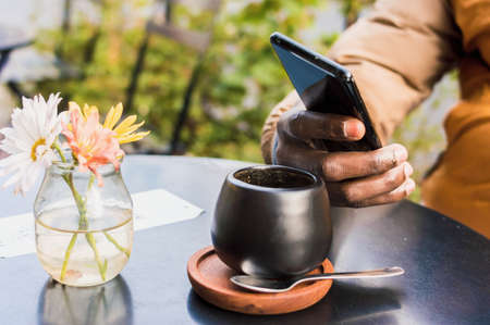 close up black male hand of unrecognizable man holding a phone on a table with a cup of coffee and a small vase in outdoors, technology concept.の写真素材