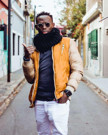 young african man with black scarf, glasses and yellow jacket standing on the street serious looking at the camera affirming with thumb up. lifestyle concept.の写真素材