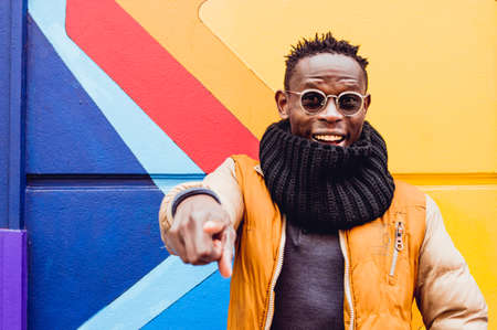 portrait of happy young african man smiling, wearing glasses and winter clothes, pointing at camera outdoors with colorful wall behind him.の写真素材