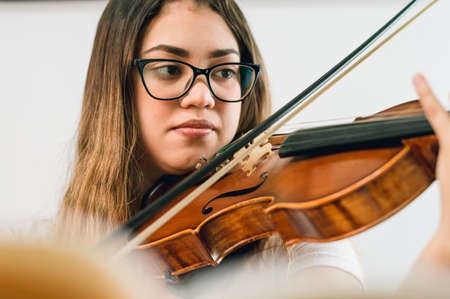 caucasian young woman with glasses, sitting on the sofa at home practicing the violin watching the...
