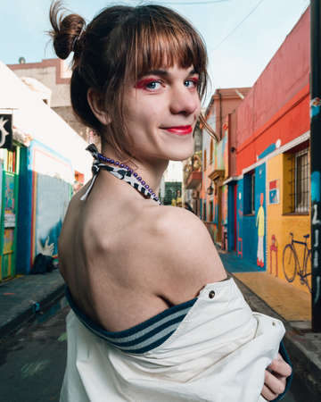 young latin caucasian transgender woman with red makeup, on the street standing sideways looking back, with a jacket half on, looking at the camera, concept of lifestyle and inclusion.の写真素材
