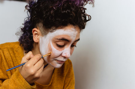 young argentinian caucasian latin woman with curlers is putting on makeup with white paint, sitting...