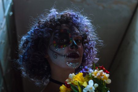 young woman with La calavera catrina makeup in the dark, looking at the camera and holding a bouquet of colorful flowers.の写真素材