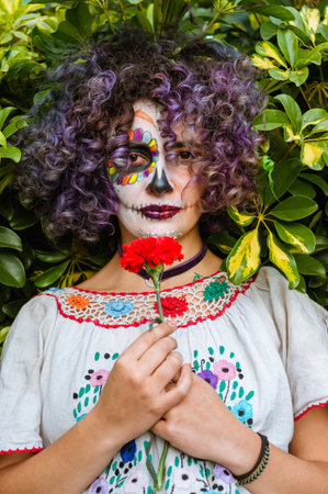vertical portrait of young latin woman outdoors holding a flower with her face painted as La Calavera Catrina and with natural green leaves in the background.の写真素材