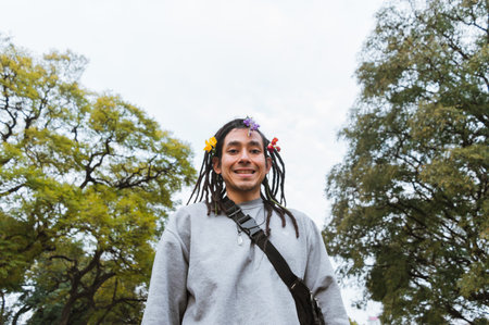 latin venezuelan young man with dreadlocks and flowers on his hair standing in the park shy and smiling looking at the camera, with trees and the sky in the backgroundの写真素材