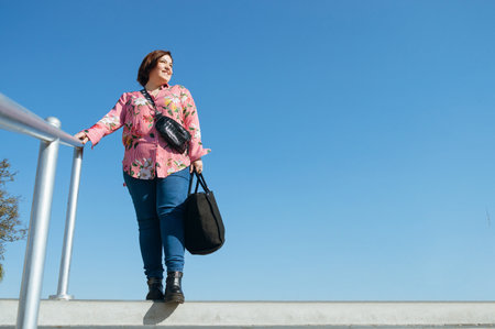young latin caucasian argentinian woman standing on the edge of stairs outdoors holding the handrail and is happy smiling turning to one side with the sky in the background and copy spaceの写真素材