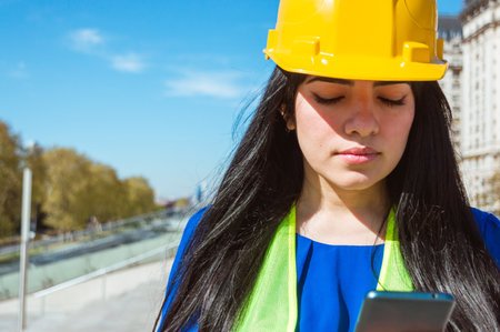 young latin caucasian venezuelan engineer woman with yellow helmet, vest and blue clothes, outdoors reading a text message with instructions from her boss to start work.の写真素材