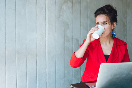 young woman in red blazer, drinking coffee while working on laptop in coffee shop, with gray wall behind her in background and copy space.の写真素材