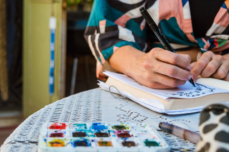 unrecognizable Caucasian female artist sitting on the terrace of the house drawing on white sheet of paper in notebook with marker and watercolors. copy space.の写真素材