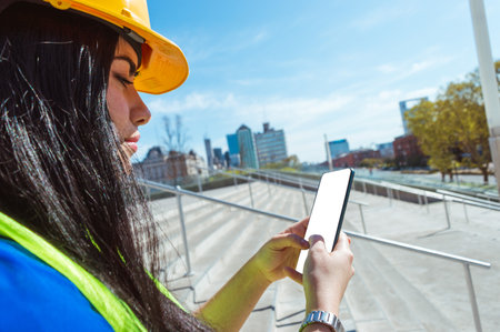 young caucasian female engineer, wearing yellow safety helmet and vest, in the street standing on the stairs texting her boss on the phone, blank screen, copy space, technology conceptの写真素材