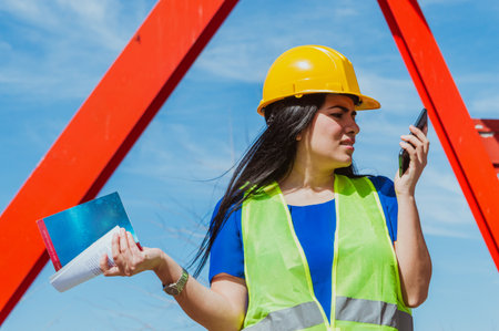 young latin caucasian woman, wearing yellow hard hat and safety vest, standing on angry construction site worker sending voice note over phone, giving instructions.の写真素材