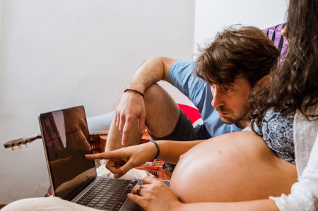 pregnant adult woman pointing at the laptop screen with her finger showing her husband the prices of diapers on the internet, they are lying on the sofa inside the house.の写真素材