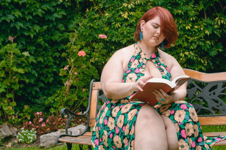 Caucasian Argentinian Latina woman plus size model reading a book sitting on a wooden bench in a garden, with a green wall in the background and copy space.の写真素材