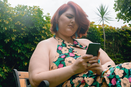 young latina caucasian argentinian woman content sitting on a bench in the park browsing the internet on her phone checking messages and social networks, with vegetation and the sky in the background.の写真素材