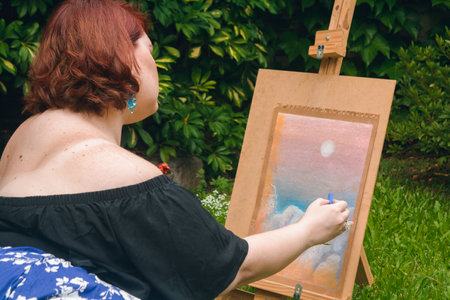 rear view of young caucasian woman with short red hair, sitting in the garden of her house, calm and alone, with a blue chalk in her hand finishing making her drawing on the easelの写真素材