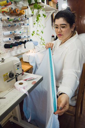 vertical image of a young argentinian woman working in her sewing workshop measuring a fabric with a tape measure, designing clothes for her clients.の写真素材