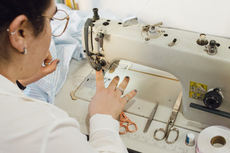 Caucasian woman threading sewing machine in her workshop, rear view