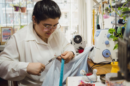 Argentinian latin young woman with glasses, standing working in her sewing workshop concentrated cutting a fabric with a scissor.の写真素材