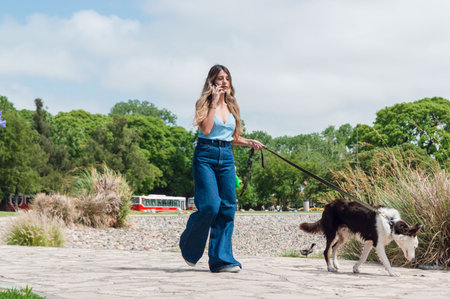 young latina colombian woman , dressed in blue and with long hair, is talking on the phone and walking with her border collie dog, walking in the park, with trees in the background and copy space.